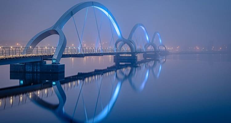 Pont piétonnier illuminé aux arches bleues se reflétant dans une rivière calme par une nuit brumeuse.