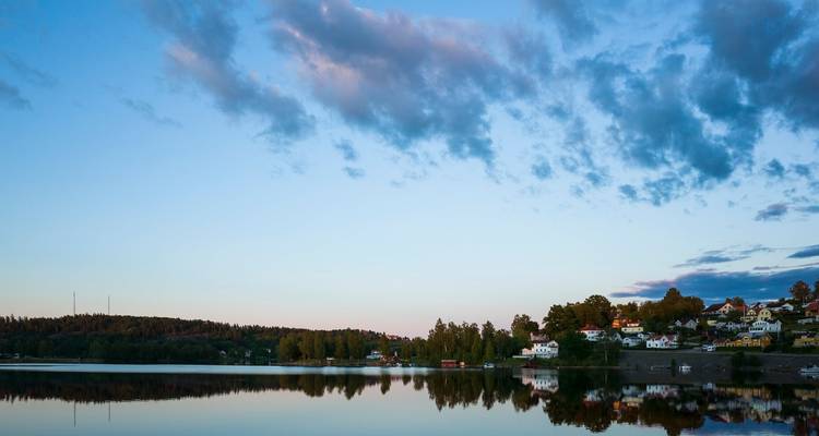 Petit village au bord du lac avec des maisons blanches reflétées dans l'eau calme du soir sous un ciel pastel.