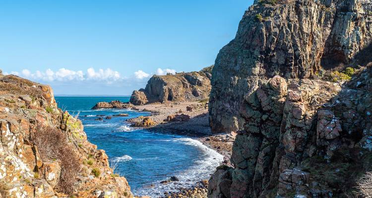 Falaises rocheuses en bord de mer et crique étroite avec des vagues qui se brisent sous un ciel bleu dégagé.