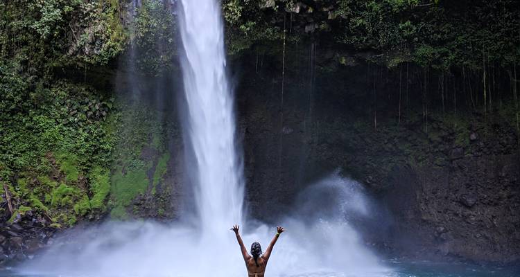 Viajero con los brazos levantados en triunfo bajo una poderosa cascada de la selva que cae en una piscina natural.