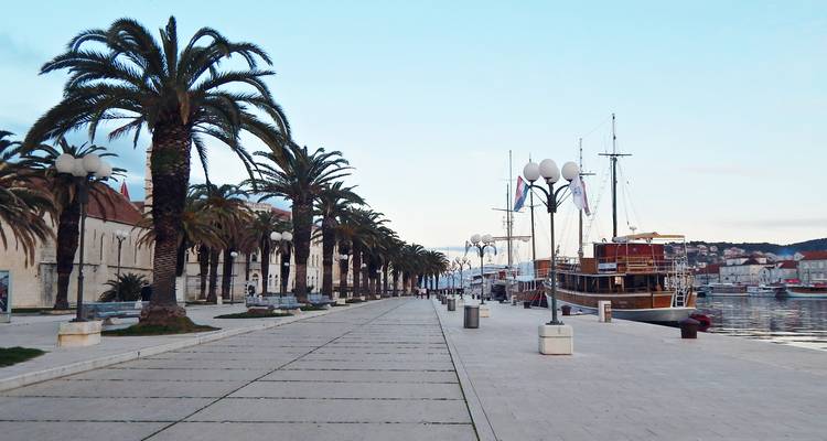 Longue promenade bordée de palmiers le long du port avec un navire en bois classique amarré au crépuscule.