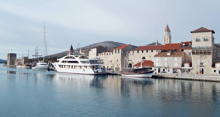 Ruhiger Hafen in Trogir, gesäumt von Steingebäuden, Booten und einem markanten Glockenturm, der sich im Wasser spiegelt.