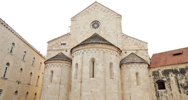 Romanische Steinkirchenfassade mit halbkreisförmigen Apsiden und einer Rosette vor weißem Himmel.