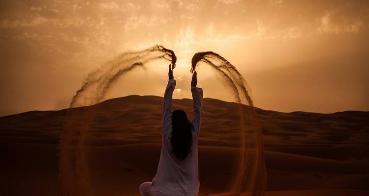 Silhouette d'une femme agenouillée dans le désert au coucher du soleil lançant des arcs de sable vers le ciel