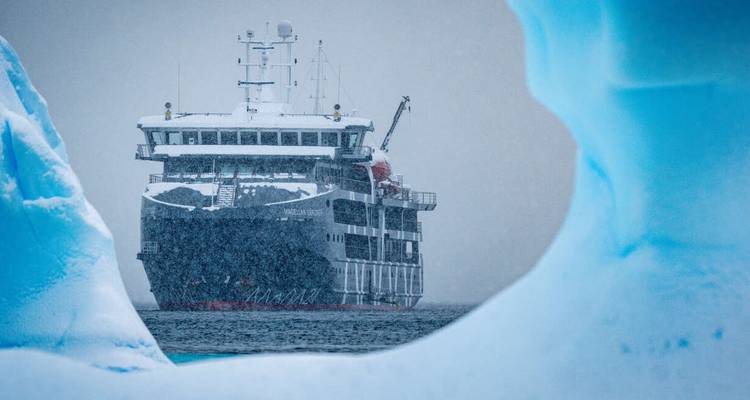 À travers une arche de glace, un navire d'expédition navigue dans les eaux antarctiques pendant une chute de neige.
