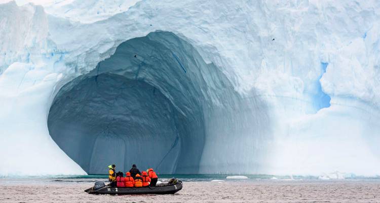 Un zodiac avec des visiteurs en vestes oranges s'approche de la vaste bouche bleue d'une grotte d'iceberg.