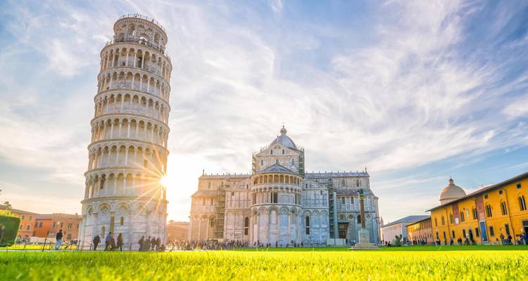 La Torre Inclinada de Pisa y la catedral brillan bajo la cálida luz del sol contra un cielo dramático con turistas dispersos en el césped verde.