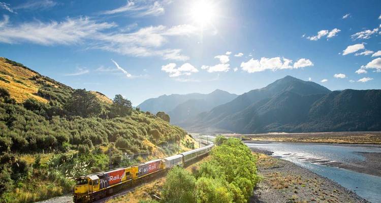 Tren panorámico de KiwiRail serpenteando a través de un exuberante valle fluvial bajo imponentes montañas bajo un sol brillante.