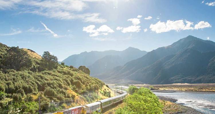 Long train de passagers glissant à travers une vallée baignée de soleil avec les imposantes montagnes de Nouvelle-Zélande.