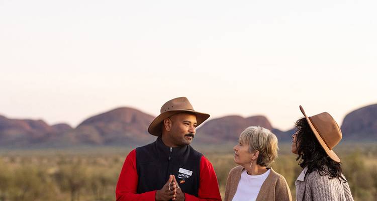 Gids en twee bezoekers met outback-hoeden in gesprek met de okerkleurige koepels van Kata Tjuta in de verte.