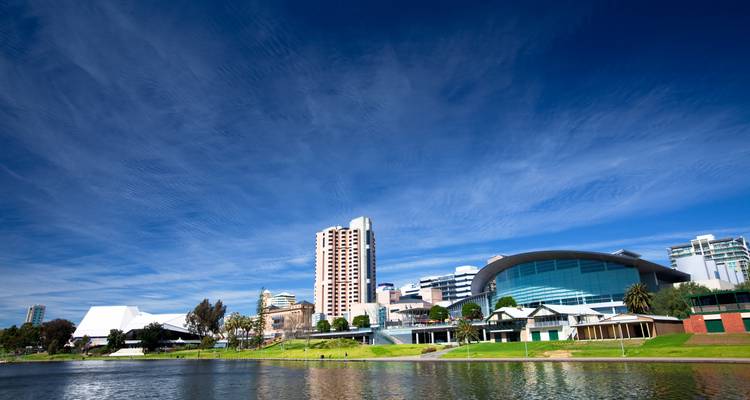 Horizonte moderno de Adelaide frente al agua con el centro de convenciones curvado, torres de gran altura y césped ribereño reflejados en el río tranquilo bajo un cielo azul profundo.