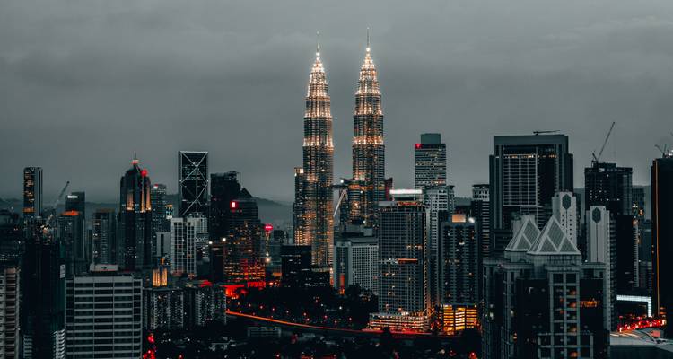 Kuala Lumpur Skyline in der Dämmerung mit leuchtenden Stadtlichtern und den Petronas Towers, die über anderen Gebäuden erstrahlen.