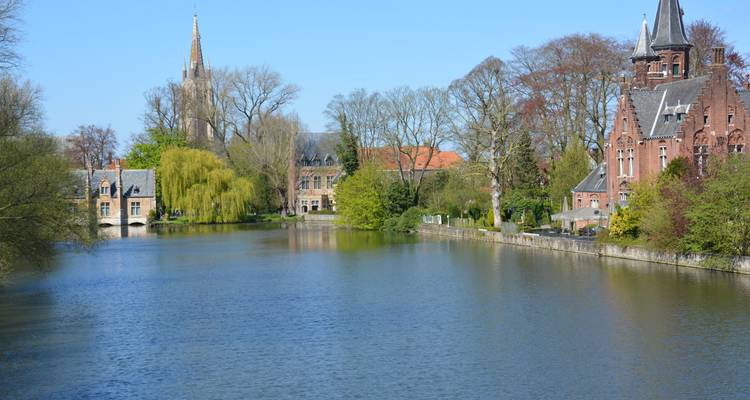 Kalme Brugse gracht omzoomd door historische bakstenen gebouwen en met bomen gevulde parken onder een heldere hemel