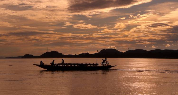 Gesilhouetteerde houten boot met passagiers die over een rustige rivier glijdt bij een vurige zonsondergang.