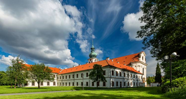 Historisches tschechisches Kloster mit roten Dächern vor dramatischen Wolken und grünen Rasenflächen.