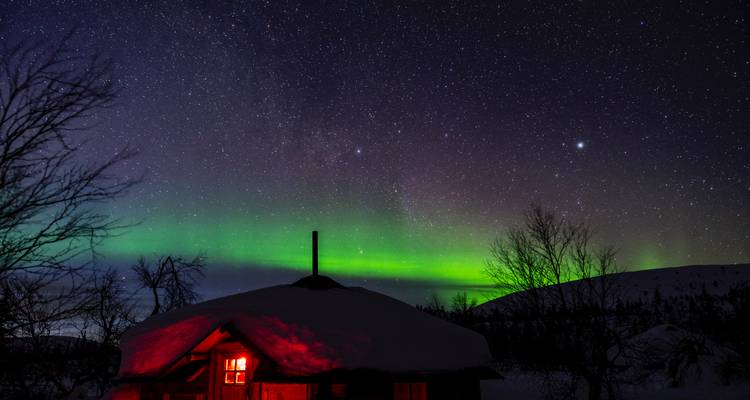 Schneebedeckte Hütte, die rot leuchtet unter einer lebendigen Aurora und dem sternenerfüllten arktischen Himmel.