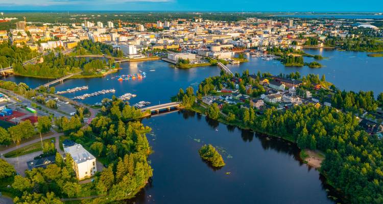 Lebendige Luftaufnahme des Stadtzentrums von Oulu, umgeben von blauen Wasserwegen und üppig grünen Inseln im Abendlicht.