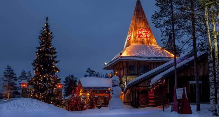 Schneebedecktes Weihnachtsmannhaus, das mit festlichen Lichtern leuchtet, neben einem geschmückten Baum in der Abenddämmerung.