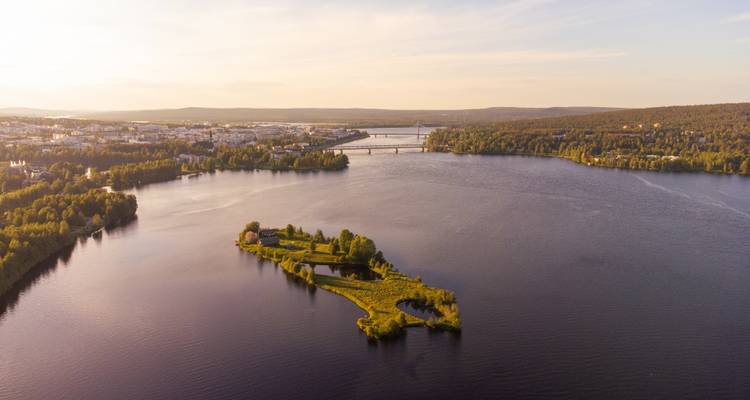 Weitläufiger See mit bewaldeter Insel und langer Brücke unter weichem Abendlicht bei Rovaniemi.