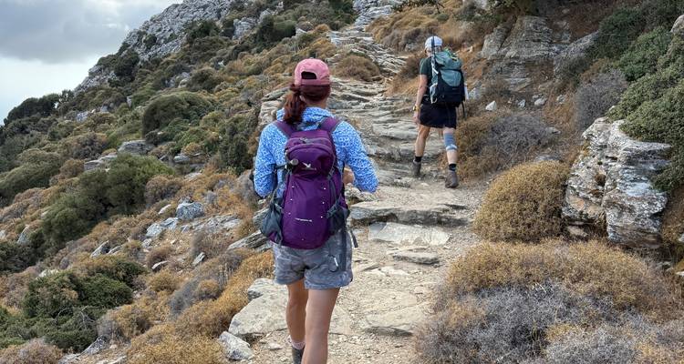 Deux randonneurs avec des sacs à dos gravissent un sentier de montagne rocailleux à travers la garrigue méditerranéenne sèche.