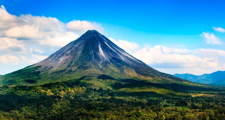 Majestic conical Arenal Volcano rises above forested plains under a bright blue sky.