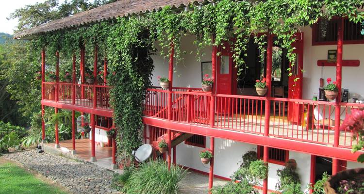 Casa tradicional colombiana estilo finca con balcones de madera roja cubiertos de exuberantes enredaderas verdes, ubicada en una ladera soleada.