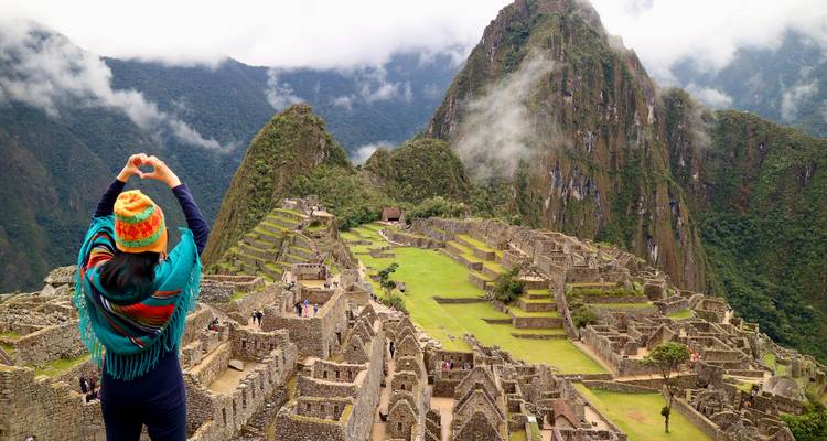 Voyageur portant un chapeau andin coloré forme un cœur avec ses mains en contemplant les ruines anciennes du Machu Picchu enveloppées de brume.