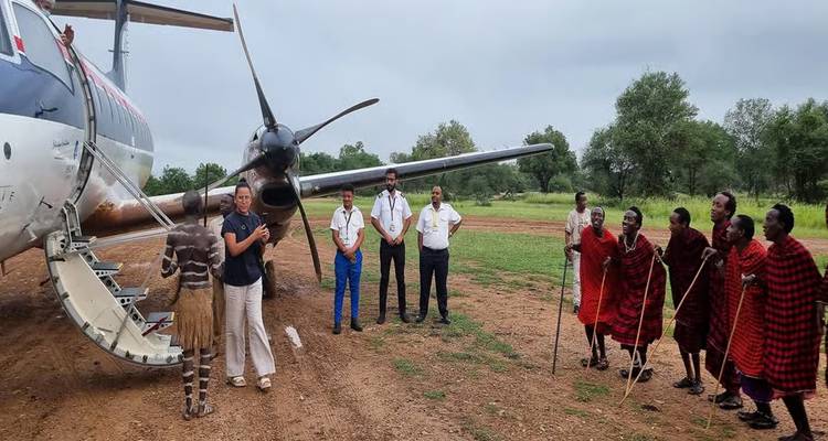 Travelers and local Maasai greet beside a small aircraft on a dirt runway.