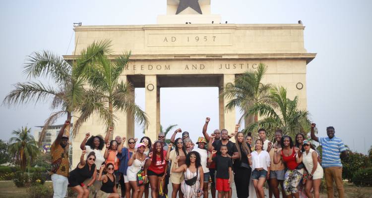 Grote tourgroep poseert voor Accra's Onafhankelijkheidsboog met palmbomen en heldere lucht.
