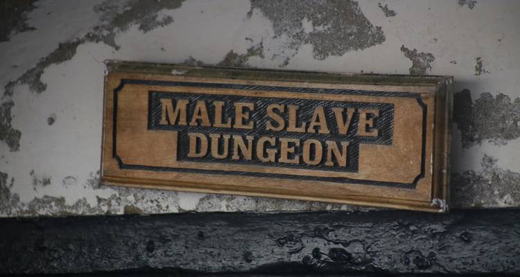 Verweerde houten plaquette met de tekst "Mannelijke Slavenkerker" op een stenen muur.