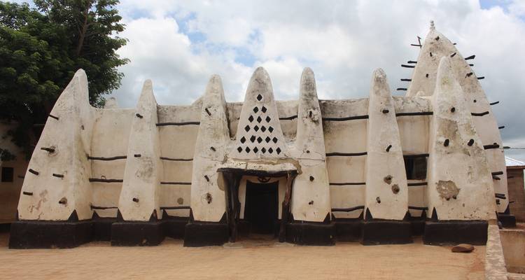 Icónica Mezquita de Larabanga de barro blanco y palos bajo un cielo parcialmente nublado.
