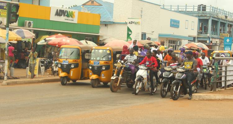 Intersección ghanesa concurrida con motocicletas, tuk-tuks y vendedores ambulantes.
