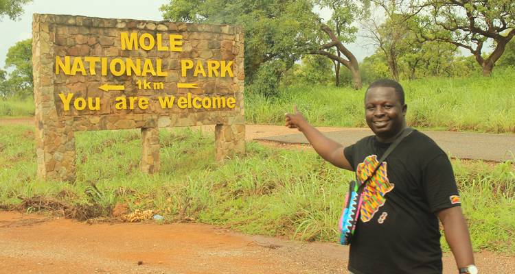 El viajero señala con orgullo el cartel de bienvenida del Parque Nacional Mole en medio de la vegetación.