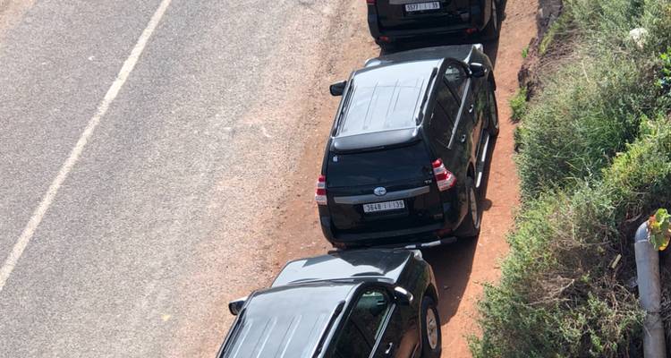 An overhead perspective shows multiple black SUVs driving along a dusty roadside verge.
