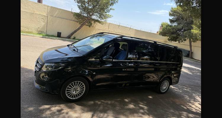 A black passenger van parked beside a tall wall topped with barbed wire under blue skies.