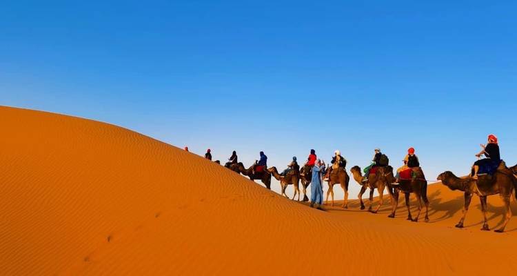 Caravane de chameaux avec cavaliers traversant de hautes dunes orangées sous un ciel désertique sans nuages.