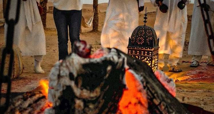 Scène nocturne de feu de camp dans le désert avec des musiciens en robes blanches jouant autour de flammes incandescentes.