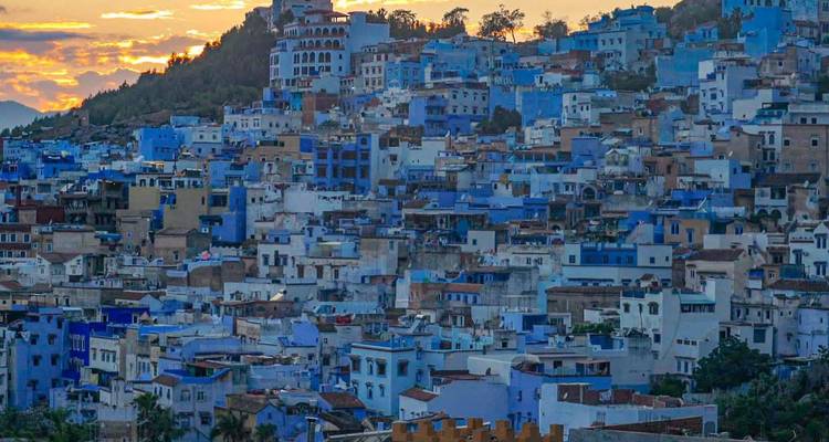 Les maisons bleues à flanc de colline de Chefchaouen au coucher du soleil avec les montagnes lointaines silhouettées contre un ciel orange.