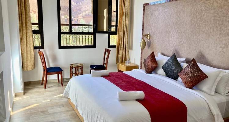 Chambre d'hôtel lumineuse avec grand lit, chemin de table rouge et vue sur la montagne par les fenêtres.