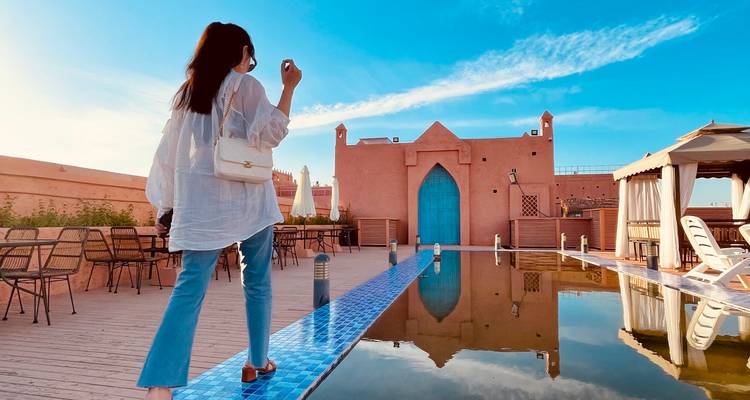 Stilvoller Reisender, der neben einem spiegelnden Becken auf einer Dachterrasse eines lehmfarbenen Riads unter blauem Himmel wandelt.
