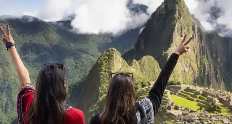 Deux voyageuses lèvent des signes de paix en faisant face au sommet emblématique et aux terrasses du Machu Picchu entourées de montagnes verdoyantes et de brume.