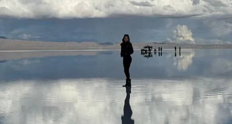 Femme seule debout sur la surface miroir du salar d'Uyuni avec des reflets de nuages spectaculaires et des silhouettes au loin.