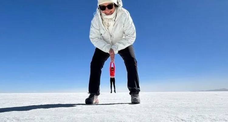 Prise de vue en perspective ludique sur le salar d'Uyuni blanc où une femme au premier plan fait semblant de tenir un petit compagnon entre ses jambes contre un ciel bleu sans nuages.