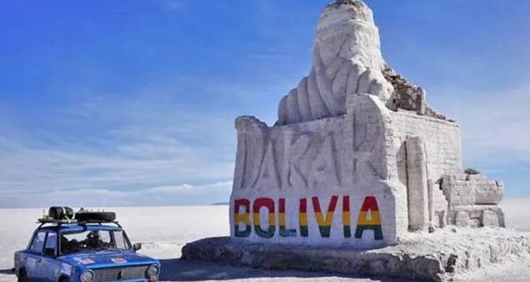 Monument de Dakar Bolivie sculpté dans le sel avec des lettres peintes aux couleurs de l'arc-en-ciel, se dressant sur la vaste étendue blanche du salar d'Uyuni sous un ciel lumineux ; une jeep bleue est garée à proximité.