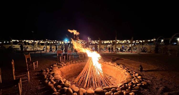 Nighttime desert camp with large bonfire surrounded by stone circle and diners under string lights.