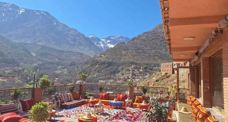 A colorful rooftop terrace with patterned rugs and low sofas overlooks snow-capped Atlas Mountains and the Imlil valley.