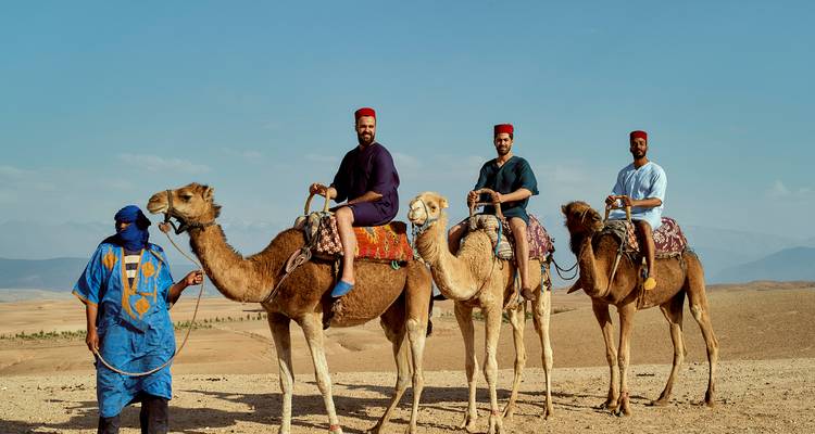 Three travelers in fez hats ride camels across a sandy plateau with distant Atlas peaks on a clear day.