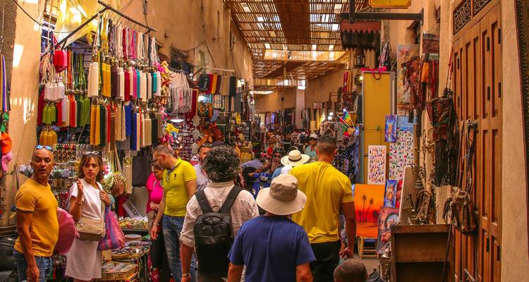 A busy covered souk packed with stalls selling textiles and trinkets as shoppers browse the narrow alley.