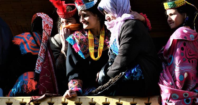 Frauen in heller ethnischer Kleidung lehnen sich über einen hölzernen Balkon, unterhalten sich und beobachten die Feierlichkeiten unten.