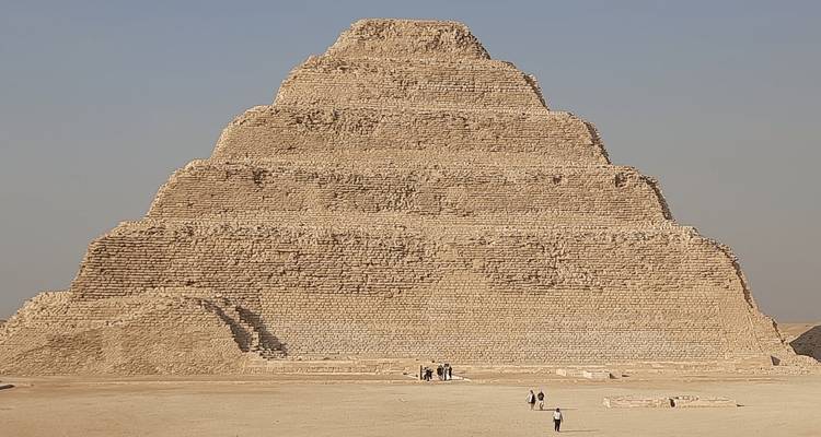 Los visitantes caminan cerca de la distintiva pirámide escalonada de Saqqara que se alza desde el desierto bajo un cielo despejado.